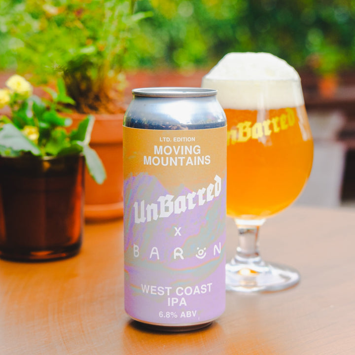 A can of UnBarred x Baron West Coast IPA sits on a wooden table next to a glass of the beer, with a blurred outdoor patio background.