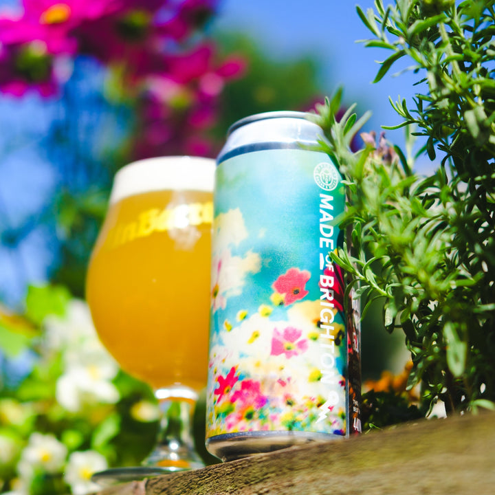 A can of Bright canned beer with a colorful floral label stands next to a filled beer glass on a wooden surface, with green plants and a blue sky in the background.