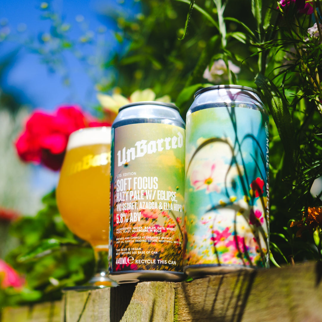 craft beer cans and a glass of hazy beer displayed outdoors on a wooden surface with flowers and greenery in the background. The center can is labeled UnBanned Soft Focus Pale Ale